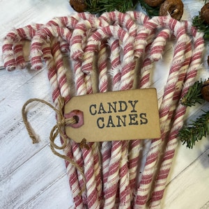 May include: A bundle of red and white striped candy canes tied together with twine. A brown tag with the words "CANDY CANES" is attached to the bundle. The candy canes are arranged on a white wooden surface with green foliage.