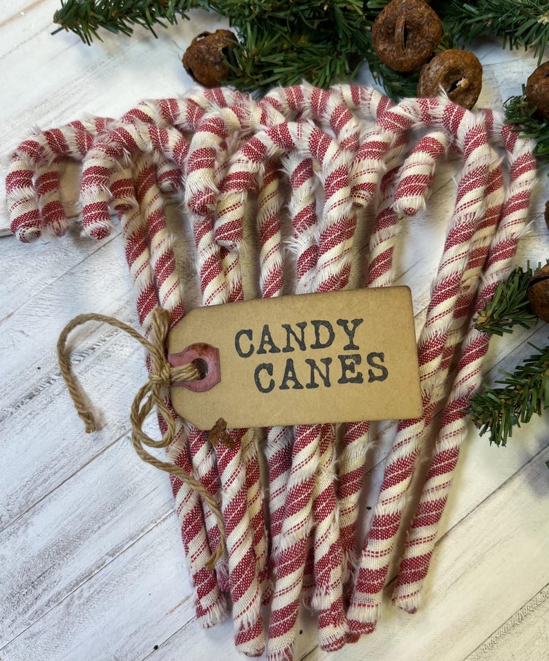 May include: A bundle of red and white striped candy canes tied together with twine. A brown tag with the words "CANDY CANES" is attached to the bundle. The candy canes are arranged on a white wooden surface with green foliage.