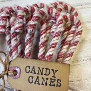 May include: A bundle of red and white striped fabric candy canes tied with twine. A brown tag with the words "CANDY CANES" in black lettering is attached to the bundle. The candy canes are arranged on a white wooden surface.