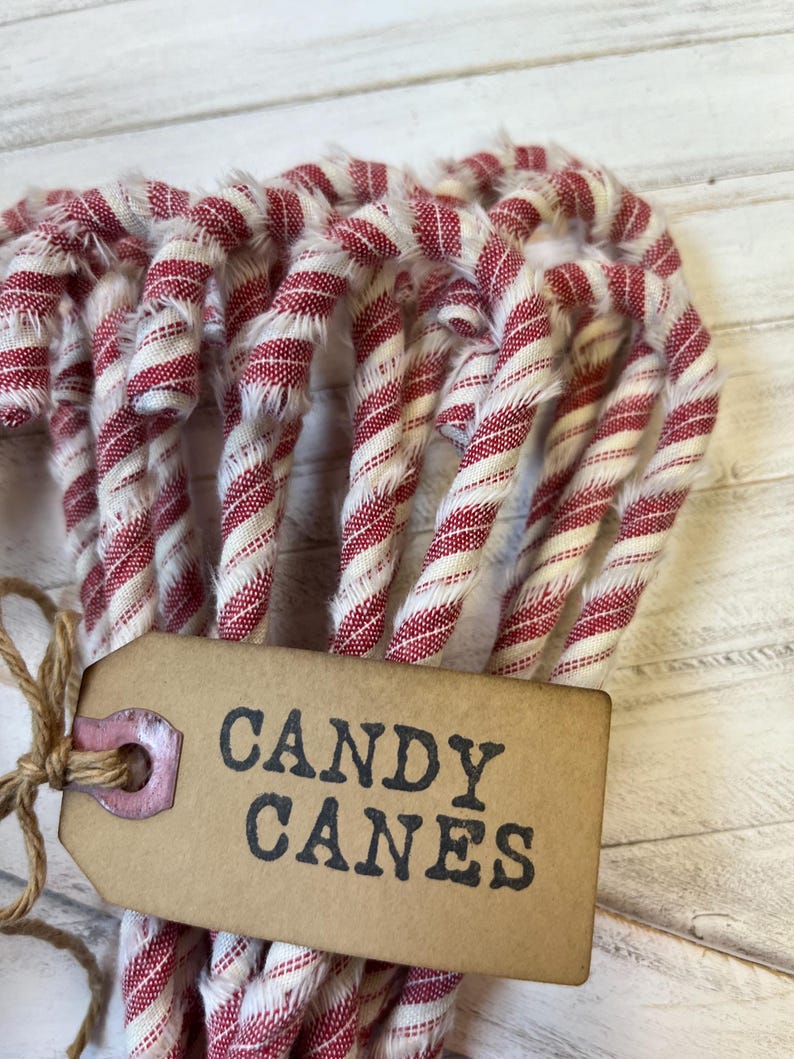 May include: A bundle of red and white striped fabric candy canes tied with twine. A brown tag with the words "CANDY CANES" in black lettering is attached to the bundle. The candy canes are arranged on a white wooden surface.