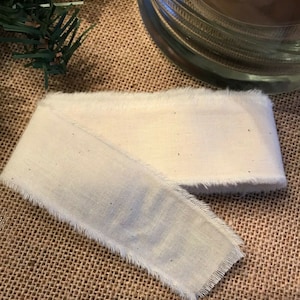 May include: A white cotton ribbon with frayed edges. The ribbon is folded in half and lying on a brown burlap surface.
