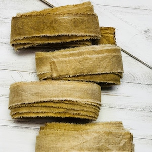May include: Three rolls of olive-gold fabric ribbon with frayed, raw edges. The ribbon has a textured, woven appearance and is coiled into loose, overlapping layers. The material displays a consistent earthy, muted yellow-green color throughout.
