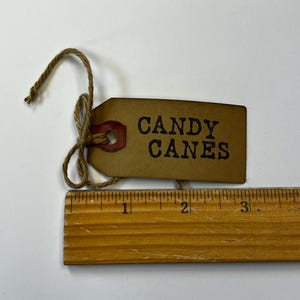 May include: A brown paper tag with the words "CANDY CANES" printed in black. The tag is tied with twine and has a red accent. A wooden ruler is placed below the tag, showing measurements in inches.