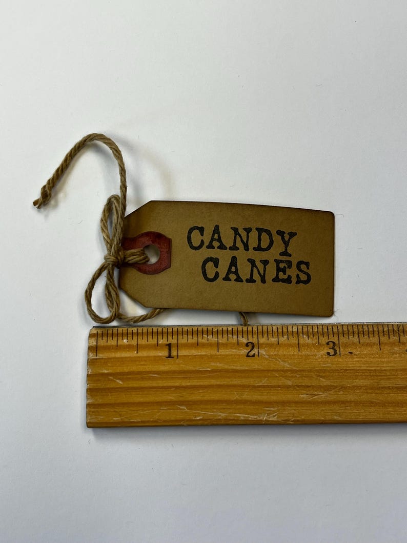 May include: A brown paper tag with the words "CANDY CANES" printed in black. The tag is tied with twine and has a red accent. A wooden ruler is placed below the tag, showing measurements in inches.