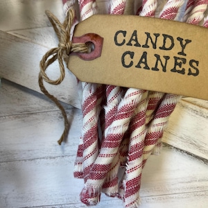 May include: A bundle of red and white striped candy canes tied with twine. A brown tag with the words "CANDY CANES" is attached to the bundle. The background is a white wooden surface.