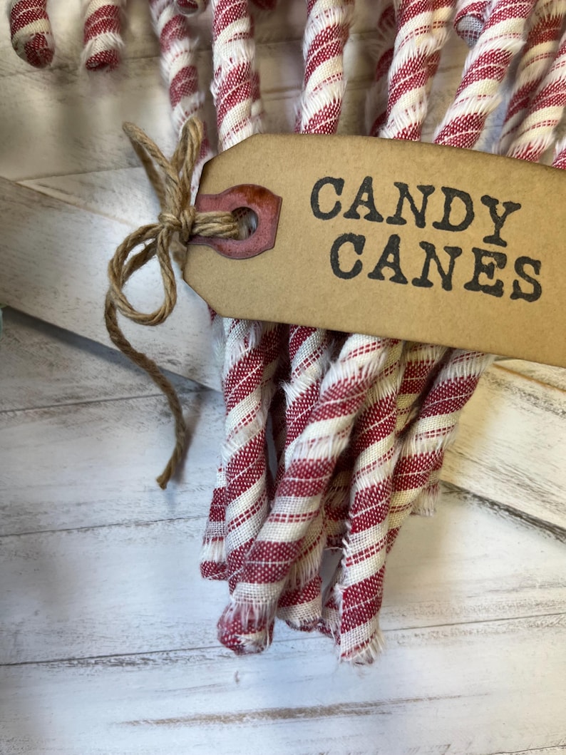 May include: A bundle of red and white striped candy canes tied with twine. A brown tag with the words "CANDY CANES" is attached to the bundle. The background is a white wooden surface.