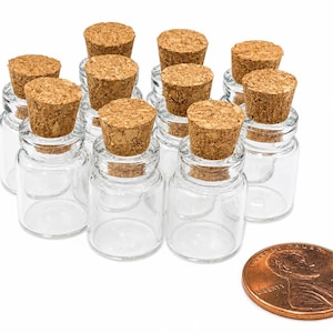 May include: A collection of ten small, clear glass bottles with cylindrical bodies and short necks, each sealed with a light brown cork stopper. A copper-colored United States penny is positioned in the foreground to provide a sense of scale.