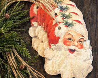 May include: A set of die-cut Santa Claus ornaments featuring a vintage illustration of a smiling Santa with a red hat, white beard, and holly sprig accents. Each ornament is attached to a natural jute twine string for hanging purposes.
