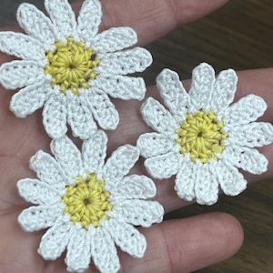 May include: Three handcrafted crochet daisy appliques featuring bright yellow centers surrounded by white petals. Each flower is constructed with fine yarn, showcasing a detailed stitch pattern that forms a circular shape with multiple distinct petals.