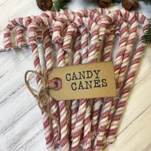 May include: A bundle of red and white striped candy canes tied together with twine. A brown tag with the words "CANDY CANES" is attached. The candy canes are arranged on a white wooden surface, with greenery and bells in the background.