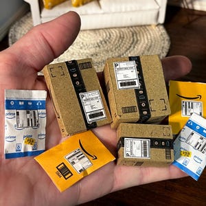 May include: Miniature cardboard boxes and envelopes, some with Amazon Prime branding and shipping labels. The boxes are brown with black tape and labels. Some envelopes are yellow and blue. A small, detailed craft project.