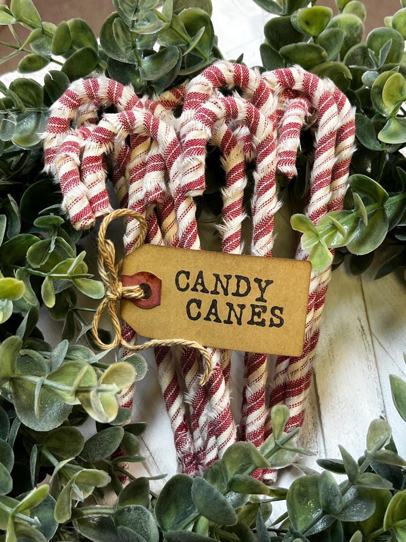 May include: A collection of red and white striped candy canes, arranged in a decorative display. A brown tag tied with twine reads "CANDY CANES". The candy canes are surrounded by green foliage, creating a festive holiday scene.