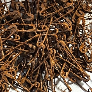 May include: A large collection of metal safety pins, each showing significant oxidation and a textured, dark brown rust coating. The pins are tangled together in a dense, overlapping pile, displaying their classic looped wire construction.
