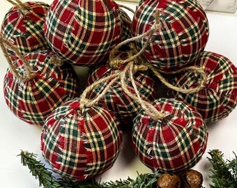 May include: A collection of plaid fabric Christmas ornaments in red, green, and cream. Each ornament is spherical, with a frayed edge design and a twine loop for hanging. The ornaments are arranged with greenery and small bells.