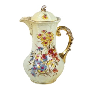Pode incluir: Um cremoso bocal de porcelana com uma alça e tampa douradas. O bocal é decorado com um padrão floral de flores amarelas, vermelhas, azuis e verdes.