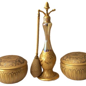 May include: A vanity set consisting of two round lidded containers and a tall perfume atomizer. Each piece features a metallic gold finish with intricate embossed scrollwork and decorative patterns. The atomizer includes a mesh bulb.