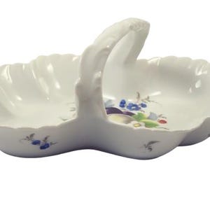 May include: White porcelain serving dish with a handle, featuring a floral design in blue, purple, and green. The dish has a scalloped edge and a handle for easy carrying. A decorative piece for serving snacks or displaying small items.