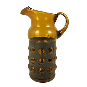 Puede incluir: Jarra de vidrio color ámbar con pico vertedor y asa. La base está envuelta en un material gris oscuro con detalles circulares color ámbar. La jarra está diseñada para servir bebidas y tiene una estética vintage.