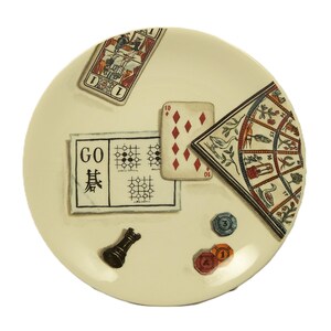 K&ouml;nnte beinhalten: Ein cremefarbener dekorativer Teller mit Illustrationen zum Thema Spiele. Er enth&auml;lt eine Spielkarte, ein Go-Brett, eine Spielkarte mit der Zahl 10, einen dekorativen F&auml;cher, eine Schachfigur und Pokerchips. Der Teller hat eine gl&auml;nzende Oberfl&auml;che.