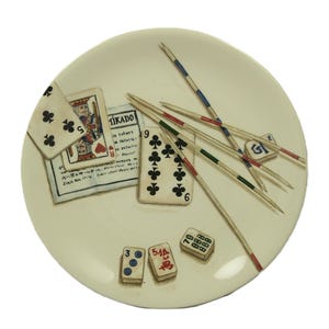 K&ouml;nnte beinhalten: Ein cremefarbener dekorativer Teller mit einem Stillleben klassischer Spiele. Der Teller enth&auml;lt Spielkarten, Mikado-St&auml;be und W&uuml;rfel. Die Spielkarten zeigen einen K&ouml;nig und eine 9 Kreuz. Die Mikado-St&auml;be sind rot, gr&uuml;n und blau.
