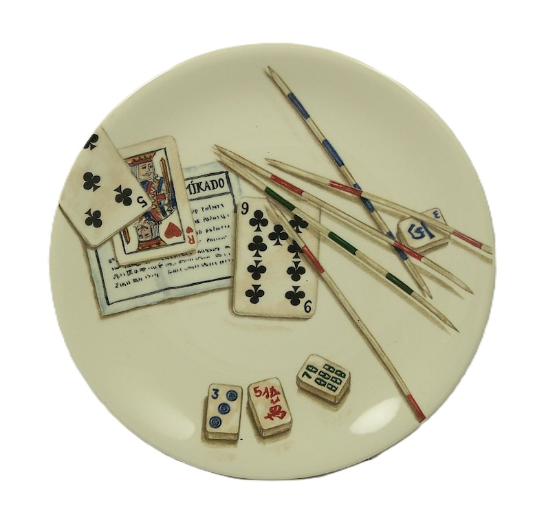 K&ouml;nnte beinhalten: Ein cremefarbener dekorativer Teller mit einem Stillleben klassischer Spiele. Der Teller enth&auml;lt Spielkarten, Mikado-St&auml;be und W&uuml;rfel. Die Spielkarten zeigen einen K&ouml;nig und eine 9 Kreuz. Die Mikado-St&auml;be sind rot, gr&uuml;n und blau.