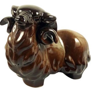 May include: A glossy, dark brown ceramic figurine of a ram with curled horns. The sculpture features a smooth, reflective glaze with lighter brown shading on the body and head, capturing the animal in a standing position with a textured coat.