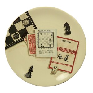 K&ouml;nnte beinhalten: Ein cremefarbener dekorativer Teller mit Illustrationen von Spielen. Der Teller zeigt ein Schachbrett, Spielkarten, ein Schachbrett, eine Schachfigur, einen Mah-Jongg-Stein und eine Mah-Jongg-Spielkarte.