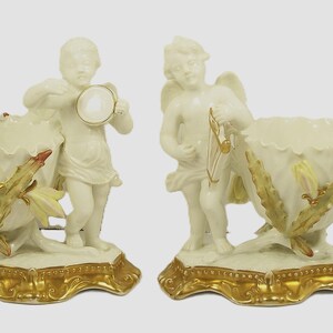 May include: Pair of ivory-colored porcelain figurines. Each features a cherub holding a musical instrument, standing beside a decorative vase. The vases have a ruffled edge and are adorned with gold and yellow leaf accents, set on a gold base.