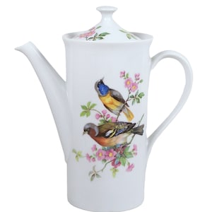 Puede incluir: Tetera de porcelana blanca con tapa y asa. La tetera tiene un diseño floral con dos pájaros en una rama.