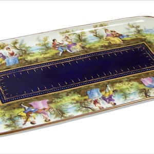 Puede incluir: Una bandeja rectangular de servir de porcelana con ribete dorado y un panel central azul y dorado. La bandeja está decorada con una escena de personas en un jardín.