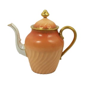 Puede incluir: Una tetera de cerámica en tonos melocotón y crema. Cuenta con un asa, un pico y un borde de tapa dorados, con un adorno dorado en la tapa. El cuerpo de la tetera presenta un diseño en espiral.