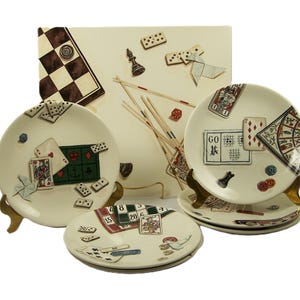 May include: A collection of cream-colored ceramic plates and a rectangular serving tray, each adorned with game-themed illustrations. The designs include playing cards, dominoes, a chessboard, and other game elements. The plates are displayed on gold-colored stands.