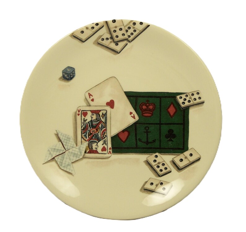 K&ouml;nnte beinhalten: Ein cremefarbener dekorativer Teller mit Spielkarten, Dominosteinen und einem W&uuml;rfel. Der Teller hat einen gr&uuml;nen Bereich mit Spielkartensymbolen. Die Spielkarten umfassen ein Herz-Ass und eine Herz-Dame.