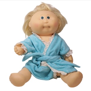 Könnte beinhalten: Eine Cabbage Patch Kid Puppe, die einen hellblauen Bademantel mit weißen Spitzenbesatz trägt. Die Puppe hat blondes Haar, blaue Augen und einen rosigen Teint.