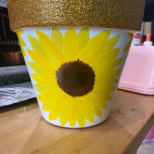 May include: A white terracotta flower pot with a gold glitter rim. The pot is decorated with a hand-painted yellow sunflower with a brown center.