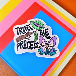 May include: A sticker with the words "Trust the Process" and illustrations of a caterpillar, chrysalis, and butterfly. The sticker is on a blue background with colorful paper layers.