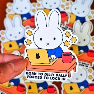 May include: A collection of stickers featuring a white bunny character with a blue shirt, working on a yellow laptop at an orange desk. The stickers have the text "BORN TO DILLY DALLY FORCED TO LOCK IN" and yellow star accents. A red mug is also on the desk.