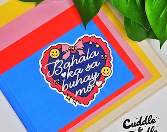 Puede incluir: Un cuaderno con una cubierta azul que presenta una pegatina en forma de corazón con el texto "Bahala ka sa buhay mo". La pegatina tiene un lazo rosa, caras sonrientes amarillas y un borde rojo. El cuaderno está rodeado de papel colorido.