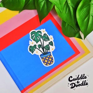 May include: A sticker featuring a potted monstera plant with large green leaves and a blue and yellow checkered pot. The sticker is on a blue notebook with a red, pink, and yellow background. The words "Cuddle & Doodle" are in black script.