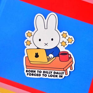 May include: A sticker featuring a white cartoon rabbit sitting at a desk with a laptop and a red mug. The rabbit is wearing a blue shirt. The text on the sticker reads "BORN TO DILLY DALLY FORCED TO LOCK IN."