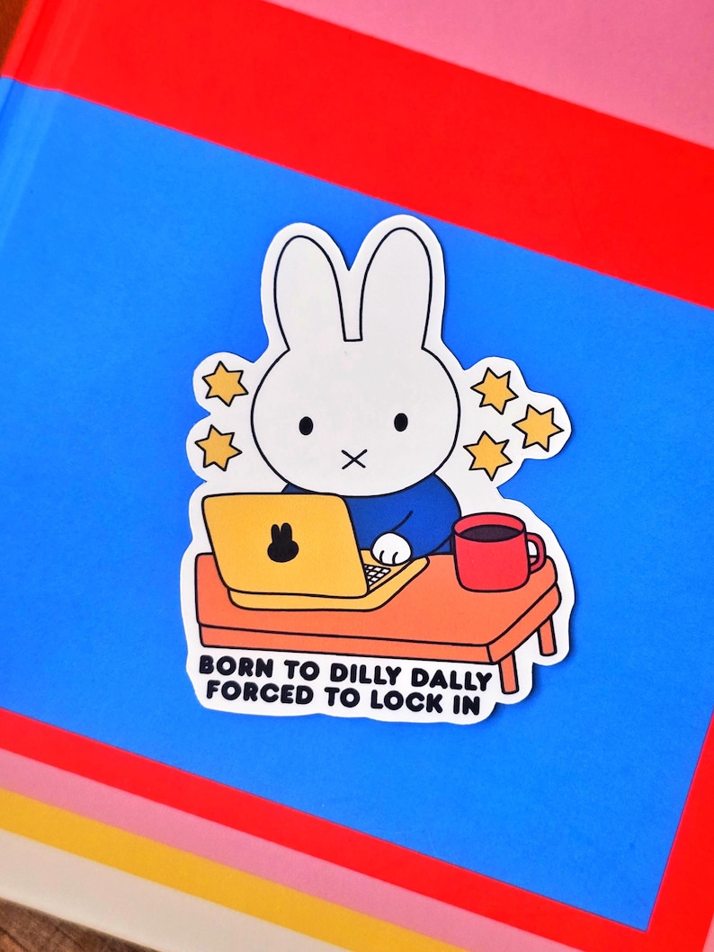 May include: A sticker featuring a white cartoon rabbit sitting at a desk with a laptop and a red mug. The rabbit is wearing a blue shirt. The text on the sticker reads "BORN TO DILLY DALLY FORCED TO LOCK IN."