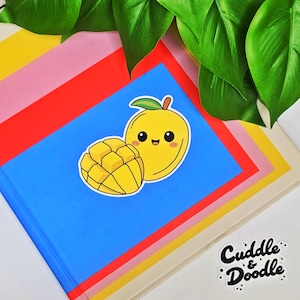 May include: A yellow mango sticker with a smiling face and a sliced mango next to it, on a blue background. The sticker is on a stack of colorful paper, with green leaves in the background. The sticker is from Cuddle & Doodle.