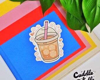 May include: A sticker featuring a cartoon iced coffee drink with ice cubes and a pink straw. The sticker is on a blue notebook with colorful paper and green leaves in the background. The sticker has the text "Cuddle & Doodle".