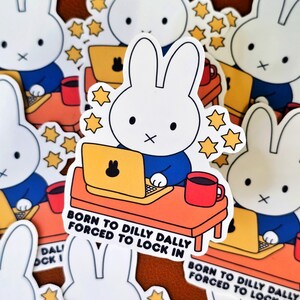 May include: White bunny stickers with a blue shirt, working on a yellow laptop at an orange desk, with a red mug. Text reads "BORN TO DILLY DALLY FORCED TO LOCK IN". Decorative star accents surround the bunny.