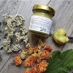May include: A small glass jar with a gold lid filled with a light green balm. The jar has a label that reads "Sweet Healing Balm" and includes instructions for use. The balm is for scrapes, bug bites, and best for skin.