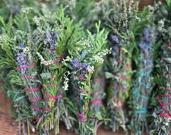 Op de afbeelding: Een rieten mand gevuld met bundels gedroogde kruiden en bloemen, gebonden met kleurrijk touw. De bundels zijn klaar om te gebruiken voor smudging of verbranding.