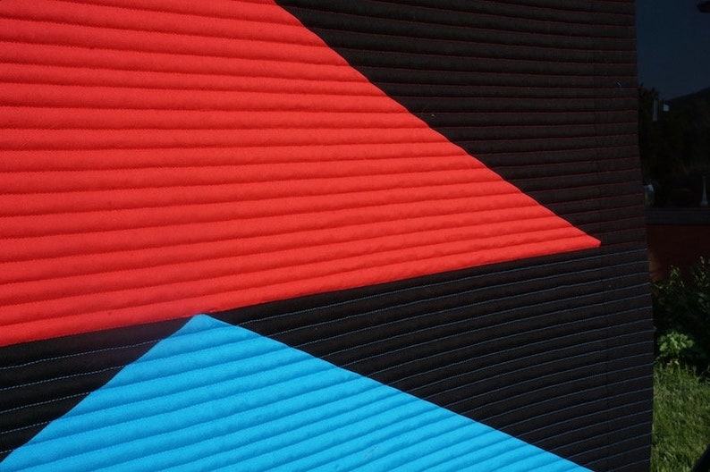 May include: A quilted wall hanging featuring three geometric shapes. The top shape is a large red triangle, the middle shape is a black triangle, and the bottom shape is a blue triangle.