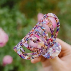May include: A large, colorful hair claw with a floral pattern. The claw is made of plastic and has a translucent appearance. The colors in the pattern include pink, blue, brown, and nude tones.