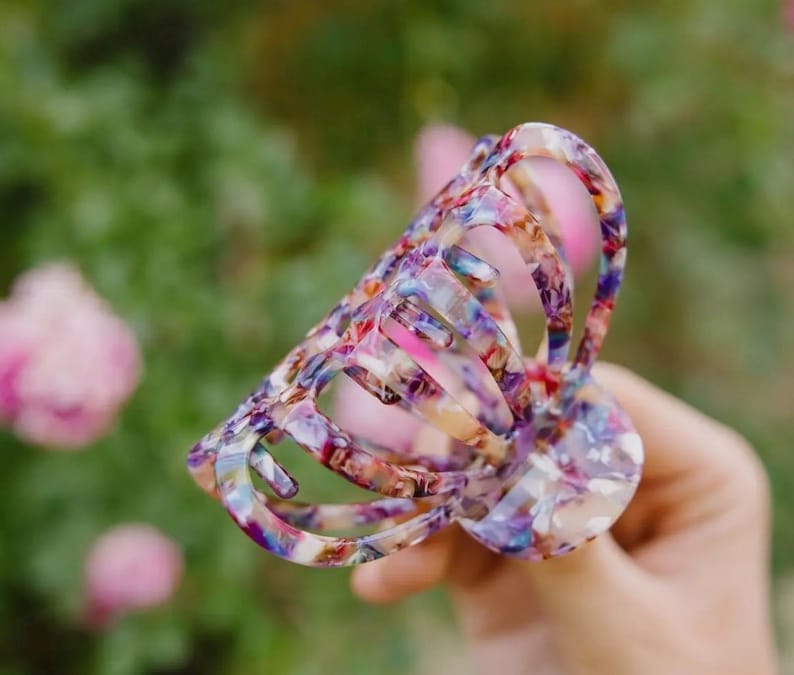 May include: A large, colorful hair claw with a floral pattern. The claw is made of plastic and has a translucent appearance. The colors in the pattern include pink, blue, brown, and nude tones.