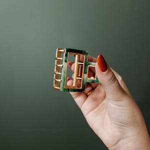 Puede incluir: Una pinza para el pelo de color verde y dorado con forma rectangular. La pinza tiene un patr&oacute;n de m&aacute;rmol y un marco de metal dorado.